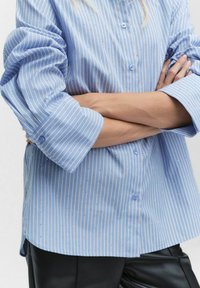 Ljust blå knapp-up skjorta med smala vita och gula ränder samt utspridda små prickar. Har uppvikt ärmar och en rundad nederkant.