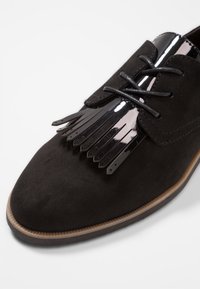 Chaussure en daim noir présentant un design de franges en cuir verni brillant sur la pointe, avec des lacets noirs et une semelle texturée.