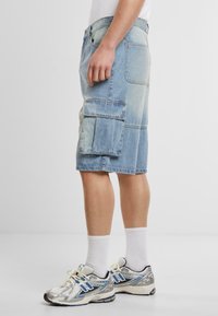 Svijetloplave denim cargo šorts hlače s opuštenim krojem, koje imaju dva velika bočna džepa i dizajn do koljena, uparene sa sivim tenisicama.
