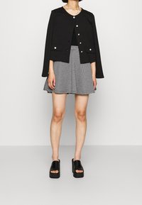 Mustat lyhythihaiset takki hopeisilla napeilla, mustan paidan päällä ja harmaa-valkoraidallinen hame, joita täydentävät mustat korokepohjaiset kengät.