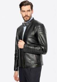 Giacca da moto in pelle nera con un design aderente, dettagli texturizzati sulle spalle, colletto alto e zip frontale. Presenta componenti hardware eleganti e minimali.