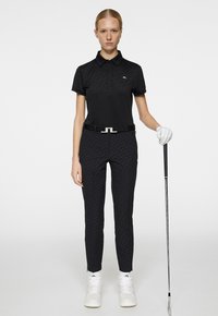 Svart pikétröja med ett strukturerat mönster, matchad med svarta mönstrade byxor. Tillbehör inkluderar ett svart bälte och vita golfskor.