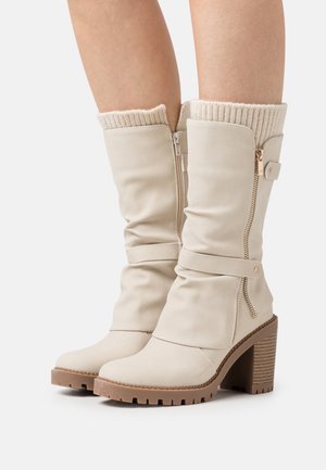 Bottes beige mi-mollet avec talons épais, fermetures éclair latérales, brides à la cheville et poignets en maille côtelée portées sur des jambes nues.
