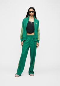 Γυναίκα που φοράει πράσινη φόρμα Adidas με κίτρινες ρίγες, μαύρο κοντό τοπ, γυαλιά ηλίου και παντόφλες, στέκεται μπροστά σε απλό λευκό φόντο.