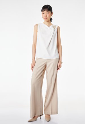 Mujer que lleva una blusa blanca sin mangas con un broche decorativo, pantalones beige de pierna ancha, tacones beige y pendientes y pulseras de aro dorados.