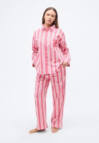 Σετ πιτζάμες με κόκκινες και λευκές ρίγες, που περιλαμβάνει πουκάμισο με κουμπιά και τσέπη στο στήθος, καθώς και φαρδιά παντελόνια, κατασκευασμένο από ελαφρύ ύφασμα.