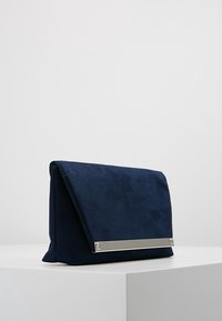 Tmavě modrá semišová kabelka s přeloženým klopou, zdobená stříbrným kovovým prvkem na přední straně a měkkou texturou.