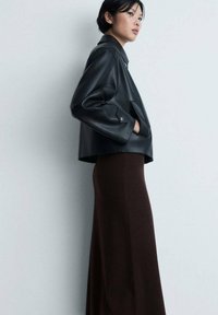 Jachetă cropped din piele neagră, cu un guler mare, asociată cu o fustă lungă și mulată de culoare maro. Textură netedă și design minimalist.
