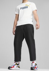 T-shirt blanc avec logo "PUMA" bleu et graphisme puma jaune, associé à un jean noir large et des baskets multicolores avec des touches rouges, bleues et blanches.