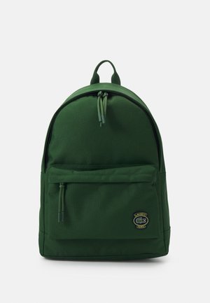 Sac à dos vert en tissu durable, présentant une forme arrondie, une poche avant zippée et un patch logo sur le bas de l'avant.
