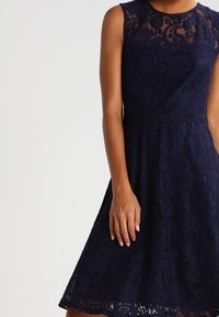 Navy lace dress with a sleeveless design, intricate floral patterns, a fitted bodice, and a flared skirt falling below the knee.