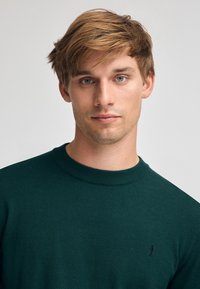 Dunkelgrüner Pullover mit rundem Ausschnitt, gerippten Bündchen und Saum. Mit kleinem, gesticktem Logo auf der linken Brust. Weiche Textur.