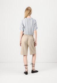 Femme aux longs cheveux blonds portant un t-shirt rayé à manches courtes, un short beige à hauteur des genoux et des ballerines noires à lacets, debout devant un fond blanc.