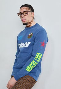 Camisa azul de manga larga con estampados gráficos, cuello redondo y letras verdes en la manga. Pantalones marrón a cuadros de corte slim. Gafas gruesas.