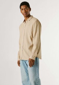 Jeune homme debout, portant une chemise beige à boutons et un jean bleu clair, sur un fond uni clair.