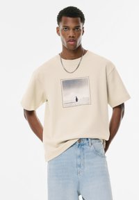 Beige bomulds T-shirt med et kvadratisk grafisk print, der viser en figur på sneklædt terræn. Kortærmet og med afslappet pasform, parret med lyseblå jeans.