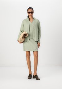 Lys grøn linsekjorte med knapper, matchende skjørt, og brune hæler. Modellen holder en beige vevd veske og har på seg solbriller.