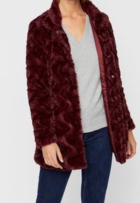 Cappotto in pelliccia sintetica bordeaux con una texture ondulata, chiusura frontale a scatto e fodera interna liscia. Indossato sopra un maglione grigio a scollo a V e jeans.