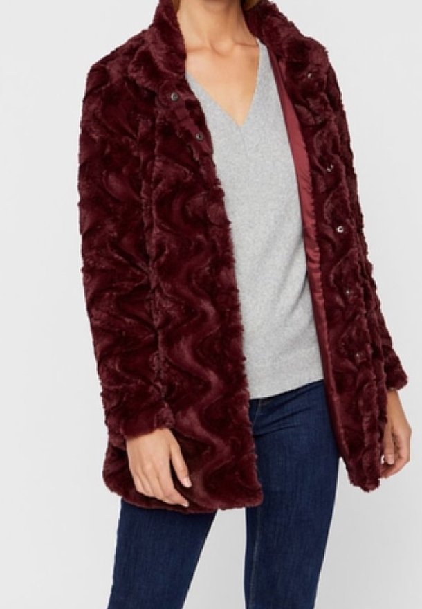 Cappotto in pelliccia sintetica bordeaux con una texture ondulata, chiusura frontale a scatto e fodera interna liscia. Indossato sopra un maglione grigio a scollo a V e jeans.