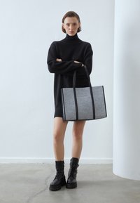 Szara torebka typu tote z czarnymi akcentami z sztucznej skóry, prostokątny kształt, dwa uchwyty i pionowe szwy. Nosi ją modelka w czarnym swetrze.