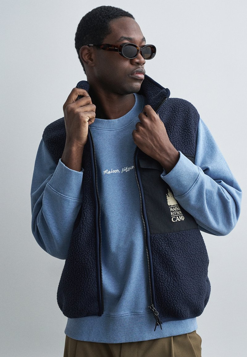 Námornícky fleece vest s nazipovaním a čiernymi textilnými akcentami. Modré mikina so slovami "Maison Kitsuné" vyšívanými bielou farbou. Textúrovaná látka.