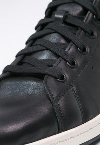 Svart lädersneaker med en slät yta, runda svarta snören, perforerade detaljer och en vit gummisula med texturerade accenter.