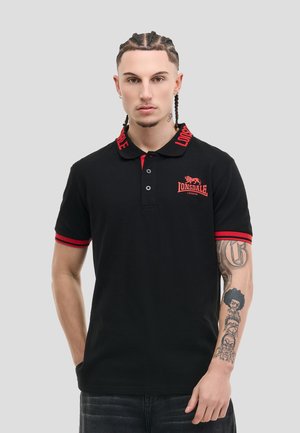 Uomo con capelli intrecciati che indossa una polo nera Lonsdale con dettagli rossi e tatuaggi sull'avambraccio destro, in piedi davanti a uno sfondo chiaro.