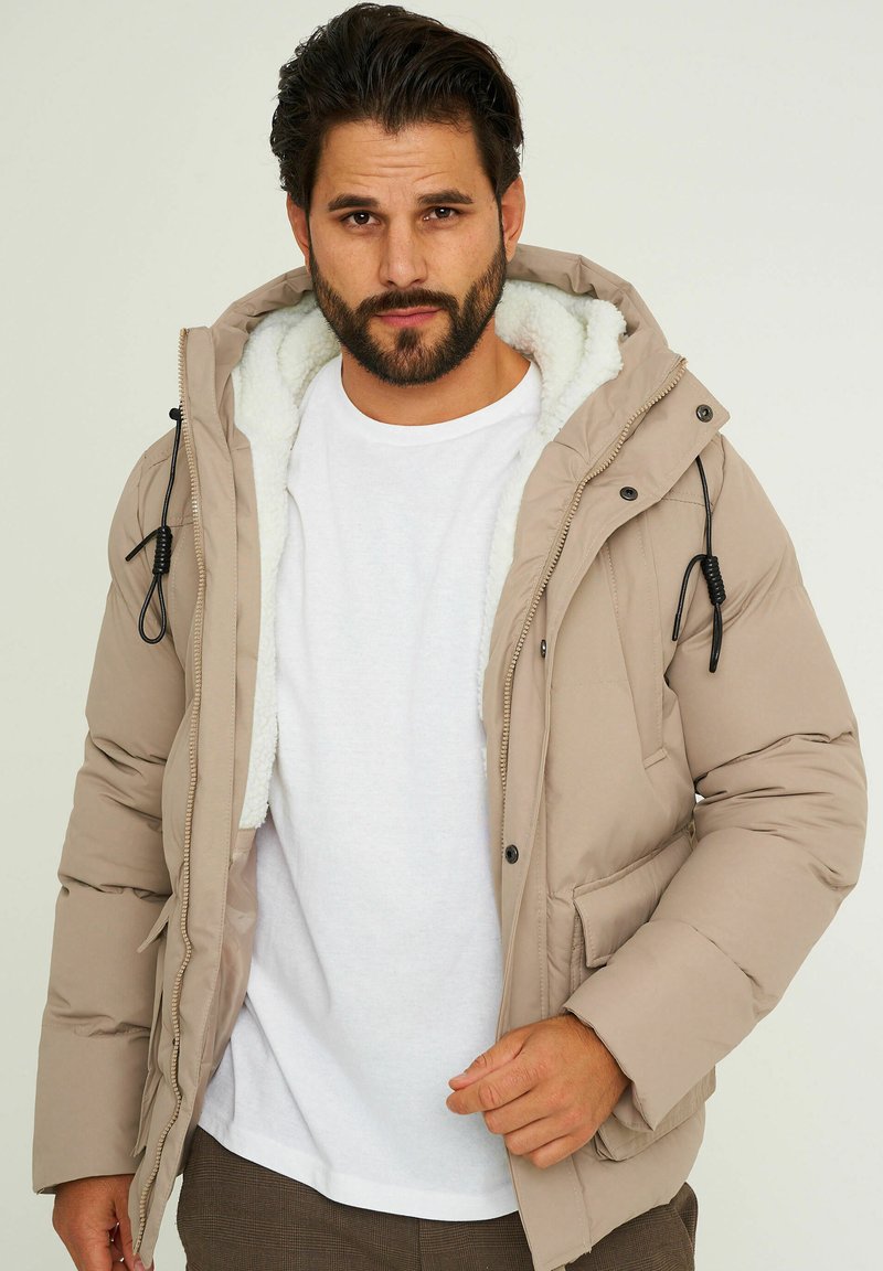 Beige gepolsterte Jacke mit weichem Innenfutter, Kordelzügen und Taschen, mit einem Reißverschluss vorne und Knopfverschluss. Darunter ein weißes T-Shirt.