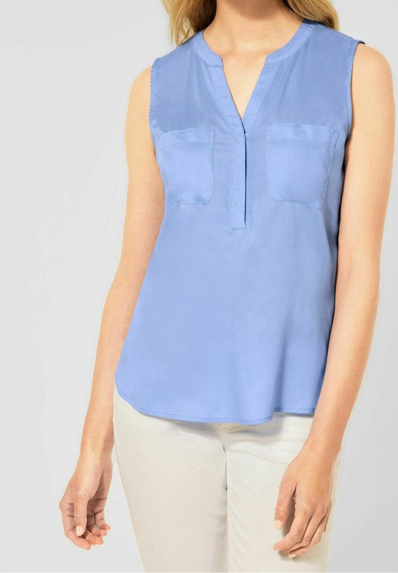 Ljusblå ärmlös topp med V-hals, två framfickor och en mjuk textur. Rundad nederkant och avslappnad passform.
