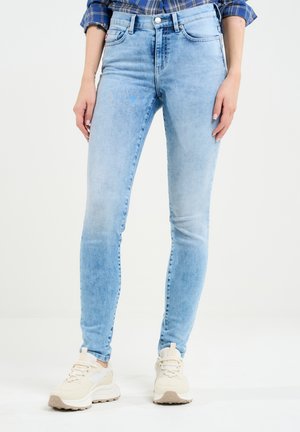 Světle modré skinny džíny s vybledlým vzhledem, pěti kapsami a zapínáním na knoflík, doplněné béžovými sportovními teniskami.