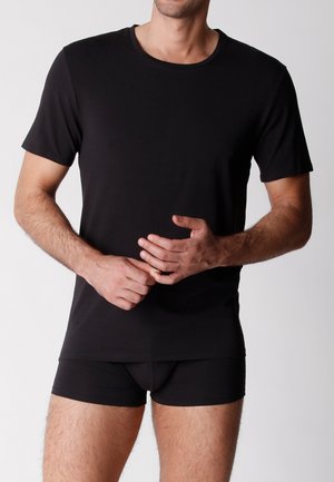 Uomo che indossa una maglietta nera semplice a maniche corte con scollo rotondo e boxer neri, in piedi davanti a uno sfondo neutro.