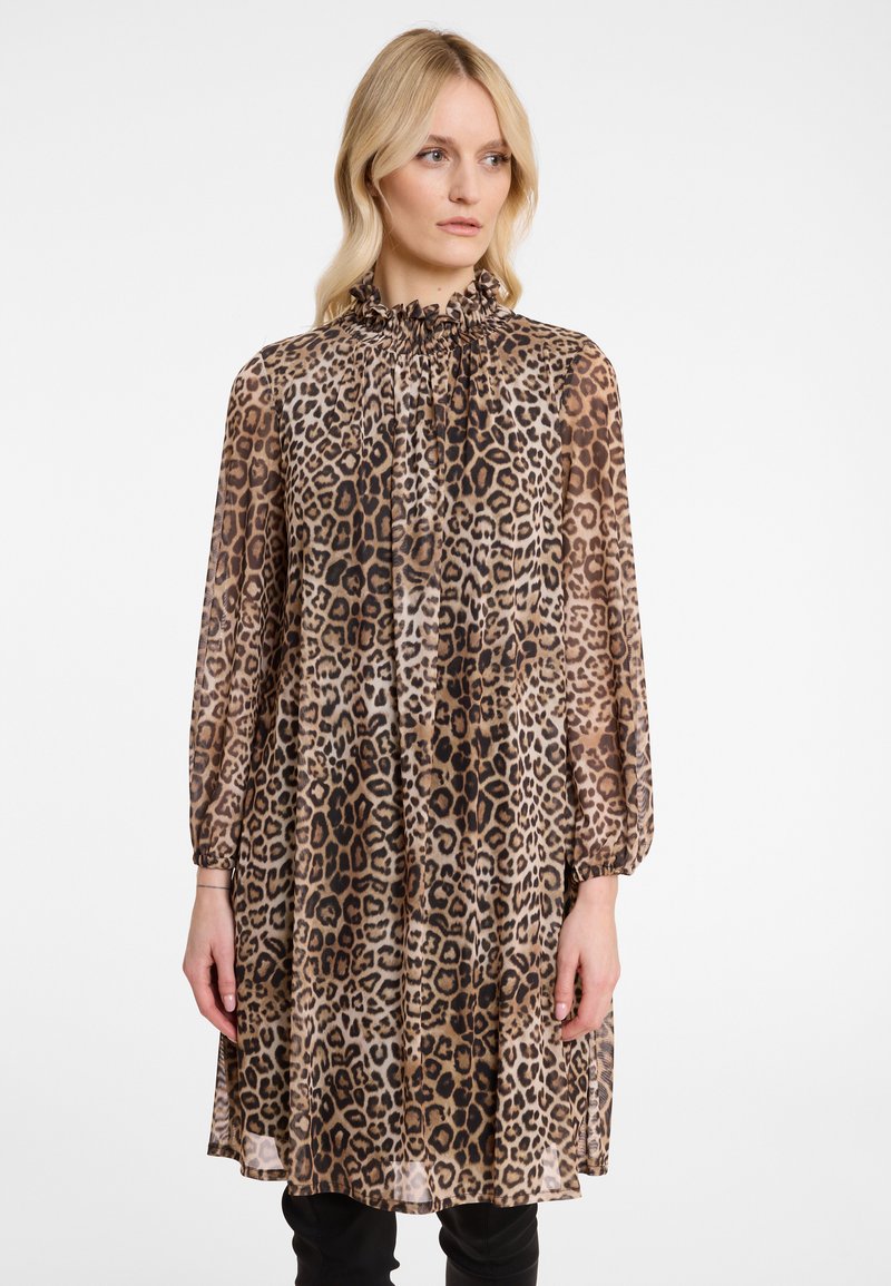 Frau in einem langärmligen, locker sitzenden Kleid mit Leopardenmuster und ruffled hohem Kragen, stehend vor einem schlichten weißen Hintergrund.