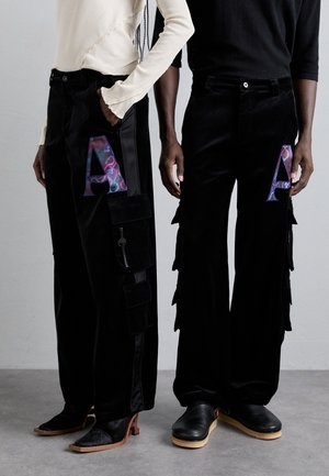 Black velvet pants with large “A” accents, featuring cargo pockets on one and layered fringes on the other. Both have unique styling details.