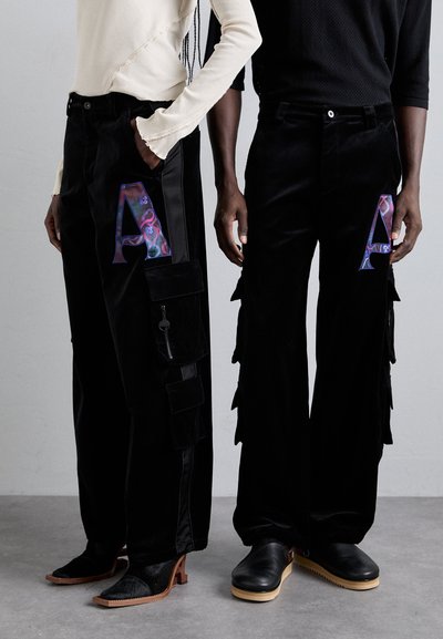 Black velvet pants with large “A” accents, featuring cargo pockets on one and layered fringes on the other. Both have unique styling details.