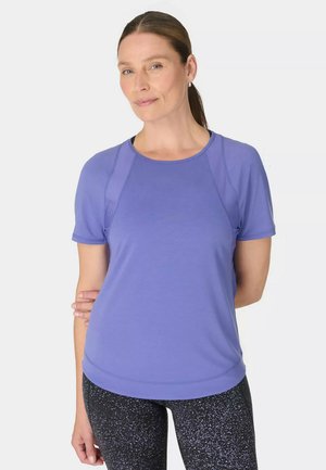 Vrouw met bruin haar in een paardenstaart, gekleed in een paarse atletische shirt met korte mouwen en zwarte legging met fijne spikkels, staand tegen een effen witte achtergrond.