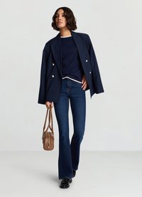 Veste croisée bleu marine avec boutons argentés, chemise bleu marine, jeans évasés foncés, et sac à main marron avec fermeture éclair. Des chaussures noires complètent la tenue.