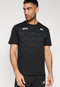 Μαύρο αθλητικό μπλουζάκι με γεωμετρικό μοτίβο υφής, κοντά μανίκια και λευκό λογότυπο UFC στο στήθος. Ομαλή υφή υφάσματος και κανονική εφαρμογή.