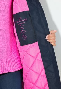 Chaqueta acolchada con tejido rosa y marino. Presenta símbolos de impermeabilidad, transpirabilidad y resistencia al viento. Bolsillo con cremallera en la manga.