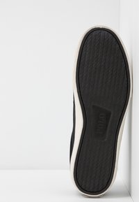 Svart skosula med texturerad yta i gummi, en vit kant och en synlig logotyp. Designen är platt med en lätt böjd form.