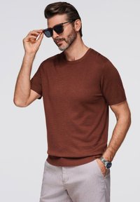 Hnědý pletený svetr s krátkým rukávem a kulatým výstřihem, doplněný o světlé kalhoty. Model má na sobě černé sluneční brýle a hodinky.