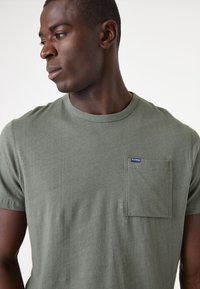 Olivgrünes Baumwoll-T-Shirt mit kurzen Ärmeln, Rundhalsausschnitt und einer Brusttasche auf der linken Seite, die ein kleines Logopatch zeigt. Glatte Textur.