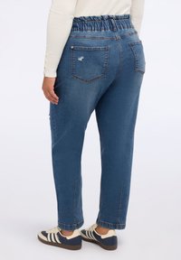 Dunkelblaue Denim-Jogginghosen mit elastischem Bund, ausgestattet mit Gesäßtaschen und einem schmal zulaufenden Bein-Design. Kombiniert mit marineblauen und cremefarbenen Turnschuhen.
