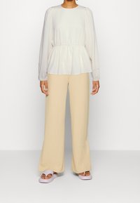 Blusa beige chiaro con vita arricciata e maniche a palloncino, abbinata a pantaloni a gamba larga crema e sandali a scivolo lavanda.