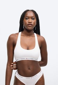 Valge puuvillane spordirinnahoidja laia ribaga, millel on "TOMMY HILFIGER" logo. Silmapaistev tekstuur, ümmargune kaelus ja laiad rihmad. Sobivad valged püksikud.