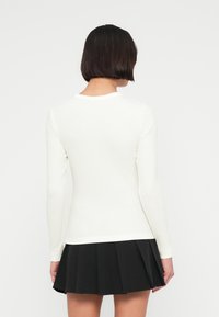 Λευκό τοπ με μακριά μανίκια, στρογγυλή λαιμόκοψη, εφαρμοστή γραμμή και ριμπ υφή. Συνδυασμένο με μαύρη πλισέ φούστα.