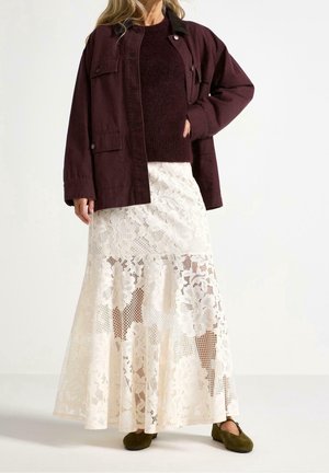 Femme portant une veste violet foncé, un pull assorti, une jupe longue blanche en dentelle fleurie et des chaussures vert olive, debout avec une main dans la poche.