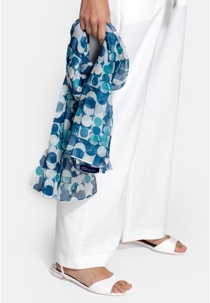 Main tenant une écharpe bleue à motifs, portant un pantalon blanc à jambes larges et des sandales plates blanches à bout ouvert sur fond blanc.