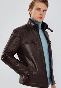 Giacca di pelle marrone con accenti trapuntati sulle spalle, tasche con zip e colletto alto, abbinata a un dolcevita azzurro chiaro sotto.