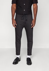 Pantalons en mélange de coton noir avec une taille à cordon de serrage, coupe fuselée et texture lisse, associés à des mocassins noirs brillants et des chaussettes blanches.