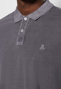 Szara koszulka polo z teksturowanej tkaniny, z dwukolorowym karczkiem zapinanym na dwa guziki oraz małym białym haftowanym logo na piersi.